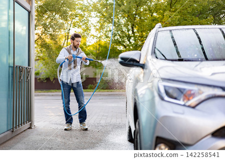 A caucasian man is actively cleaning his silver car using a high-pressure water jet at an outdoor self-service car wash station A caucasian man is actively cleaning his silver car using a high-pressure water jet at an outdoor self-service car wash station 142258541