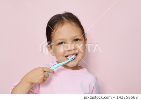Happy little girl brushing her teeth with a toothbrush, smiling at the camera against a pink studio background Happy little girl brushing her teeth with a toothbrush, smiling at the camera against a pink studio background 142258669