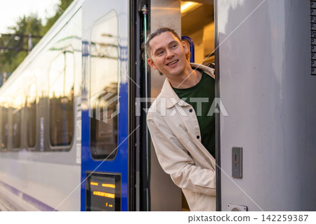 Cheerful mid adult man looking out of passenger train door and smiling during stop. Concept of travel joy rail tourism vacation emotions and destination arrival. Cheerful mid adult man looking out of passenger train door and smiling during stop. Concept of travel joy rail tourism vacation emotions and destination arrival. 142259387