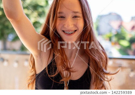 Red haired Gen Z teenager with braces laughs joyfully with arm raised while holding smartphone. Concept of authentic teen confidence and infectious playful happiness in everyday lifestyle moment. Red haired Gen Z teenager with braces laughs joyfully with arm raised while holding smartphone. Concept of authentic teen confidence and infectious playful happiness in everyday lifestyle moment. 142259389