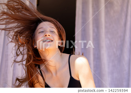 Red haired teenage girl with braces laughs joyfully while dancing with hair blowing on sunny balcony. Concept of infectious happiness and carefree youthful energy expressed through dance. Red haired teenage girl with braces laughs joyfully while dancing with hair blowing on sunny balcony. Concept of infectious happiness and carefree youthful energy expressed through dance. 142259394