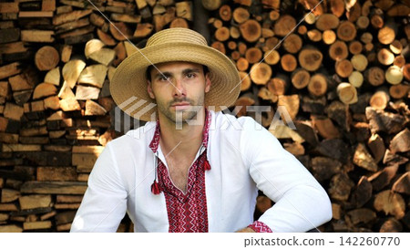 Portrait of ukrainian guy in straw hat against the background of woodshed. Young man wearing an embroidered shirt looking into camera outdoor. Concept of cultural heritage and ethnic roots Portrait of ukrainian guy in straw hat against the background of woodshed. Young man wearing an embroidered shirt looking into camera outdoor. Concept of cultural heritage and ethnic roots 142260770