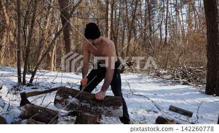 Shirtless man sawing a log in sunny winter forest. Strong guy working with saw in snowy woodland. The scene highlights human effort and outdoor activity in cold conditions. Slow motion Shirtless man sawing a log in sunny winter forest. Strong guy working with saw in snowy woodland. The scene highlights human effort and outdoor activity in cold conditions. Slow motion 142260778
