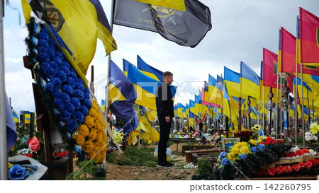 Mourning man standing among countless graves of ukrainian soldiers with national banners pay tribute to fallen family member in Kharkiv. Scene reflect personal sorrow loss caused by russian aggression Mourning man standing among countless graves of ukrainian soldiers with national banners pay tribute to fallen family member in Kharkiv. Scene reflect personal sorrow loss caused by russian aggression 142260795