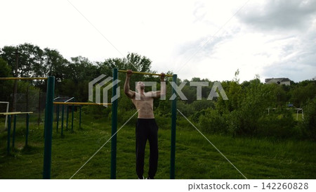 Muscular athlete doing pull ups on horizontal bar at sports ground. Young shirtless sportsman exercising at nature. Strong sporty man training outdoor. Concept of healthy and active lifestyle. Slow mo Muscular athlete doing pull ups on horizontal bar at sports ground. Young shirtless sportsman exercising at nature. Strong sporty man training outdoor. Concept of healthy and active lifestyle. Slow mo 142260828