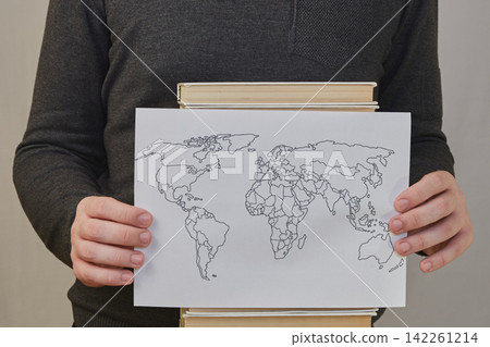 A person's hands holding a black-and-white world map against a stack of books. A person's hands holding a black-and-white world map against a stack of books. 142261214