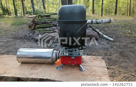 A gray camping pot with a lid sits on a red portable gas stove, connected to a silver canister on a wooden surface, with a blurred forest background. A gray camping pot with a lid sits on a red portable gas stove, connected to a silver canister on a wooden surface, with a blurred forest background. 142262751