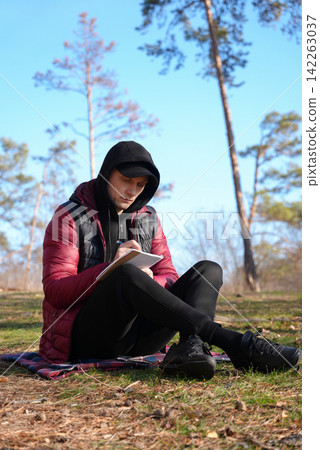Young man writing in notebook while sitting in forest Young man writing in notebook while sitting in forest 142263037