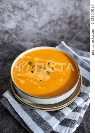 Bowl of creamy pumpkin soup topped with pumpkin seeds. Rich orange puree is served in rustic bowl and decorated with crunchy seeds creating an appetizing autumn meal presentation. Concept of seasonal Bowl of creamy pumpkin soup topped with pumpkin seeds. Rich orange puree is served in rustic bowl and decorated with crunchy seeds creating an appetizing autumn meal presentation. Concept of seasonal 142263056