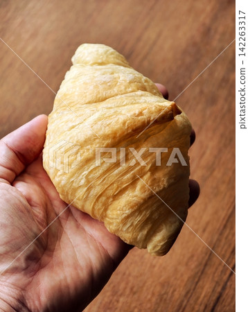Hand holding a freshly baked golden butter croissant over a wooden table. 142263317