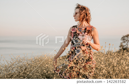Woman flowers sunset, stylish woman in floral dress stands in field of wildflowers during golden hour near the ocean shore Woman flowers sunset, stylish woman in floral dress stands in field of wildflowers during golden hour near the ocean shore 142263582