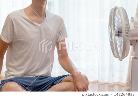 A man sitting on the floor of a room, enjoying the breeze from a fan. A man sitting on the floor of a room, enjoying the breeze from a fan. 142264982