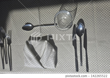 Elegant Table Setting With Folded Napkin, Silverware and Glass Cast in Strong Sunlight Shadows Elegant Table Setting With Folded Napkin, Silverware and Glass Cast in Strong Sunlight Shadows 142265522