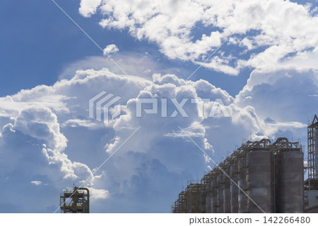 Rows of cylindrical storage tanks and pipe structures at a petrochemical plant silhouetted against a dramatic blue sky with towering cumulonimbus clouds creating a striking contrast between nature and Rows of cylindrical storage tanks and pipe structures at a petrochemical plant silhouetted against a dramatic blue sky with towering cumulonimbus clouds creating a striking contrast between nature and 142266480
