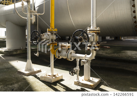Close-up of handwheel valves and yellow pipeline system installed at the base of a large insulated storage tank casting dramatic shadows on the concrete ground at an industrial plant. Close-up of handwheel valves and yellow pipeline system installed at the base of a large insulated storage tank casting dramatic shadows on the concrete ground at an industrial plant. 142266591