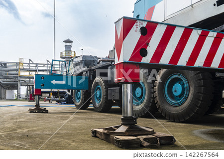 A large mobile crane with red striped body and blue wheels deployed with outriggers extended at a petrochemical plant site in an industrial heavy equipment close-up shot. 142267564