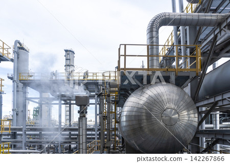 A large spherical pressure vessel with steam venting amid pipe racks and yellow walkways at a petrochemical plant under an overcast sky showcasing the grand scale of industrial energy infrastructure. A large spherical pressure vessel with steam venting amid pipe racks and yellow walkways at a petrochemical plant under an overcast sky showcasing the grand scale of industrial energy infrastructure. 142267866