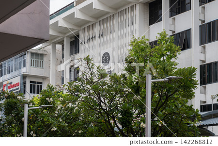 The mid-century facade of Thammasat University Faculty of Political Science Building (Tha Phra Chan) stands behind a lush green tree canopy. The white concrete structure showcases a geometric grid of The mid-century facade of Thammasat University Faculty of Political Science Building (Tha Phra Chan) stands behind a lush green tree canopy. The white concrete structure showcases a geometric grid of 142268812
