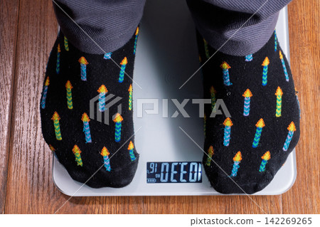 Top view of feet in socks standing on a digital weighing scale. Person measuring body weight of 103 kg on wooden floor. Obesity and diet concept Top view of feet in socks standing on a digital weighing scale. Person measuring body weight of 103 kg on wooden floor. Obesity and diet concept 142269265