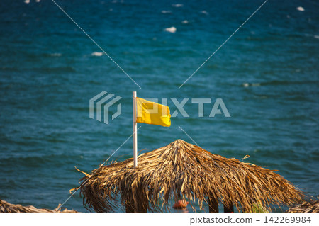 Thatched Beach Umbrella with Yellow Flag Above Blue Sea Thatched Beach Umbrella with Yellow Flag Above Blue Sea 142269984