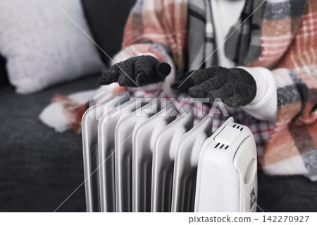 Person in gloves and blanket warming hands over an electric heater at home. Facing a heating problem, high energy costs and cold weather snap during the winter energy crisis. Person in gloves and blanket warming hands over an electric heater at home. Facing a heating problem, high energy costs and cold weather snap during the winter energy crisis. 142270927