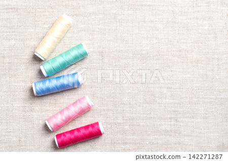 Polyester sewing thread spools randomly distributed on linen fabric, from above, with copy space. Multi colored vibrant polyester sewing thread wound onto paper rolls in a crisscross pattern. Photo Polyester sewing thread spools randomly distributed on linen fabric, from above, with copy space. Multi colored vibrant polyester sewing thread wound onto paper rolls in a crisscross pattern. Photo 142271287