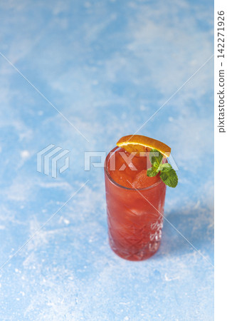Highball glass of fresh made blood orange margarita with slices of blood orange, ice cubes and mint on the blue concrete table surface. Highball glass of fresh made blood orange margarita with slices of blood orange, ice cubes and mint on the blue concrete table surface. 142271926