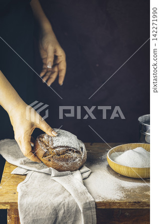 Baker's hands with a bread. Photo with high contrast. Woman hand with rye bread bread on dark wooden table with flour Baker's hands with a bread. Photo with high contrast. Woman hand with rye bread bread on dark wooden table with flour 142271990