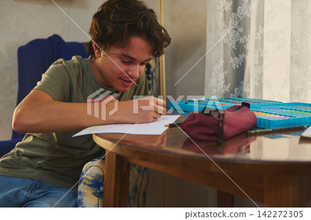 Teen boy concentrates on drawing at a home table, using colored pencils and an art set for homework and creative practice, relaxed and focused in a cozy interior with sunlight and curtains. Teen boy concentrates on drawing at a home table, using colored pencils and an art set for homework and creative practice, relaxed and focused in a cozy interior with sunlight and curtains. 142272305