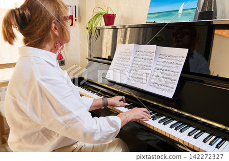 A woman practices classical piano at home during a piano class, reading sheet music with a metronome nearby. Warm interior light and focused posture convey concentration, learning and musical A woman practices classical piano at home during a piano class, reading sheet music with a metronome nearby. Warm interior light and focused posture convey concentration, learning and musical 142272327