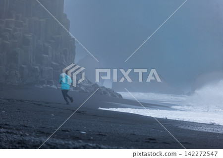 Runner in blue jacket on black sand beach Iceland with misty basalt cliffs Runner in blue jacket on black sand beach Iceland with misty basalt cliffs 142272547