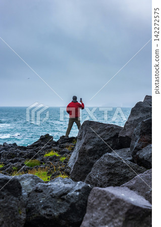 Person in Red Jacket Taking Photo on Rocky Icelandic Coastline Person in Red Jacket Taking Photo on Rocky Icelandic Coastline 142272575