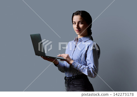 Young female student holding laptop computer and looking at camera on grey background 142273178