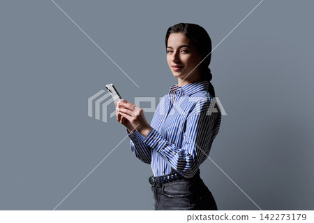 Young woman holding smartphone in half turn and looking at camera over grey background Young woman holding smartphone in half turn and looking at camera over grey background 142273179