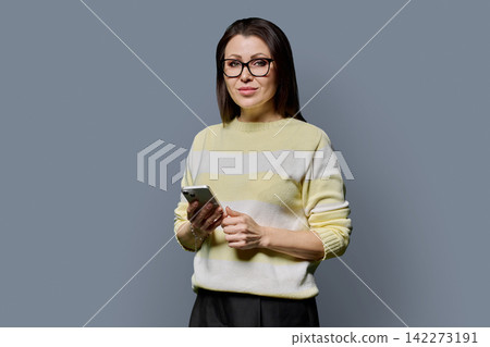 Portrait of mature woman in glasses holding smartphone on grey background Portrait of mature woman in glasses holding smartphone on grey background 142273191