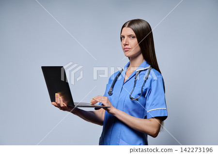 Smiling female nurse holding open laptop looking at camera isolated against grey background 142273196
