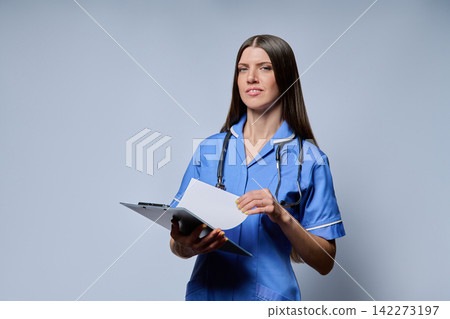 Smiling female nurse holding clipboard looking at camera against grey background Smiling female nurse holding clipboard looking at camera against grey background 142273197