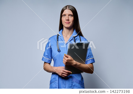 Smiling female nurse holding clipboard with patient records standing against grey background Smiling female nurse holding clipboard with patient records standing against grey background 142273198