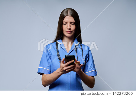 Female nurse using smartphone looking at screen against grey studio background Female nurse using smartphone looking at screen against grey studio background 142273200
