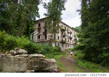 Abandoned multi-story building in an overgrown lush forest, Akarmara, Abkhazia. Tourist with a backpack walking on a dirt path towards a decaying historic structure. Abandoned multi-story building in an overgrown lush forest, Akarmara, Abkhazia. Tourist with a backpack walking on a dirt path towards a decaying historic structure. 142273584