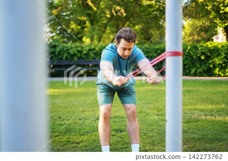 man doing an outdoor fitness workout in the park for his health. He pulls a resistance band during a strength training exercise. man doing an outdoor fitness workout in the park for his health. He pulls a resistance band during a strength training exercise. 142273762