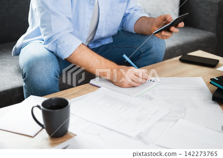 Man calculating household expenses and utility bills at home. He faces financial pressure from inflation and economic crisis, writing a budget to manage rising heating costs. Man calculating household expenses and utility bills at home. He faces financial pressure from inflation and economic crisis, writing a budget to manage rising heating costs. 142273765