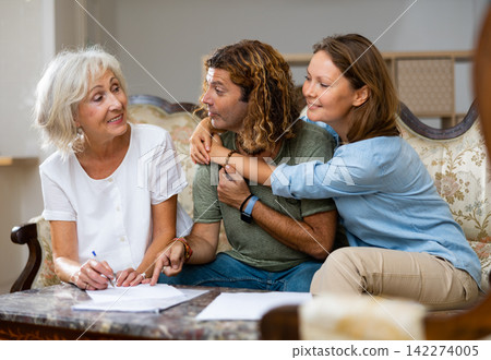 Elderly woman at table in home room filling up documents with family Elderly woman at table in home room filling up documents with family 142274005