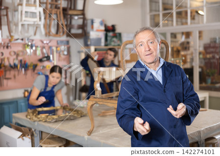 Professional elderly craftsman restorer in uniform looking at camera while working in repair shop Professional elderly craftsman restorer in uniform looking at camera while working in repair shop 142274101