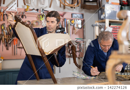Middle-aged male restorer removes old cloth seat covering from antique wooden chair. Middle-aged male restorer removes old cloth seat covering from antique wooden chair. 142274126