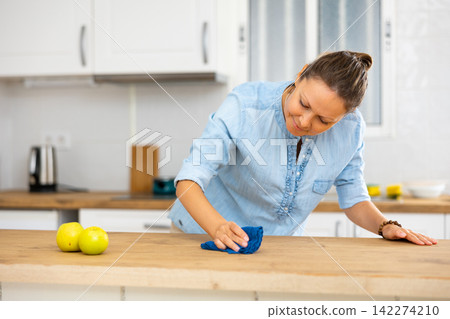 Female in casual blouse rubbing kitchen table with a rag before cooking in the cuisine Female in casual blouse rubbing kitchen table with a rag before cooking in the cuisine 142274210