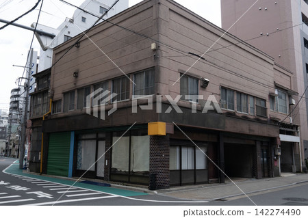 一棟復古的昭和時代建築,帶有招牌,矗立在街角,外觀像是一家老式商店兼住宅,百葉窗緊閉。 一棟復古的昭和時代建築,帶有招牌,矗立在街角,外觀像是一家老式商店兼住宅,百葉窗緊閉。 142274490