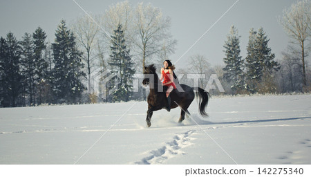 Woman riding black horse galloping in winter snow field Woman riding black horse galloping in winter snow field 142275340