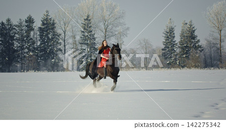 Woman riding black horse through winter snow Woman riding black horse through winter snow 142275342