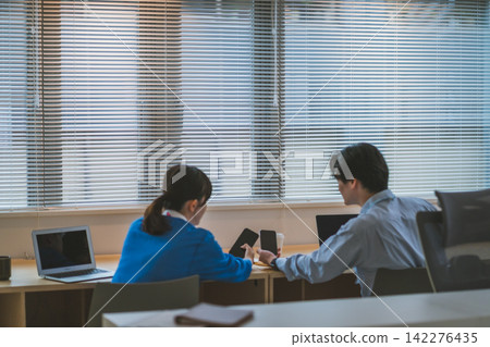 辦公室裡,男女同事互相展示智慧型手機交流。 辦公室裡,男女同事互相展示智慧型手機交流。 142276435